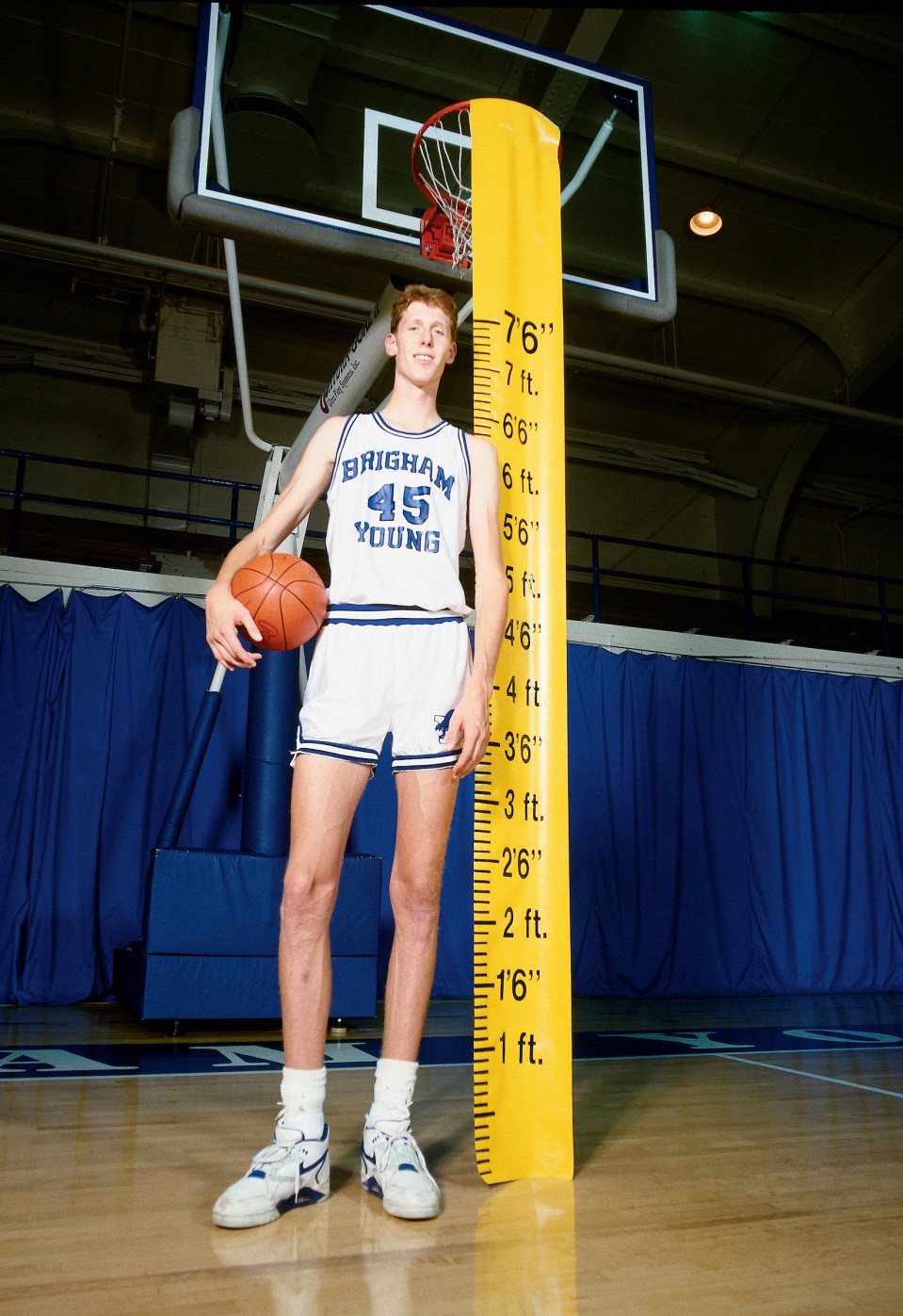 Photo of man in basketball uniform, on basketball court, standing beside ruler reading 7'6"