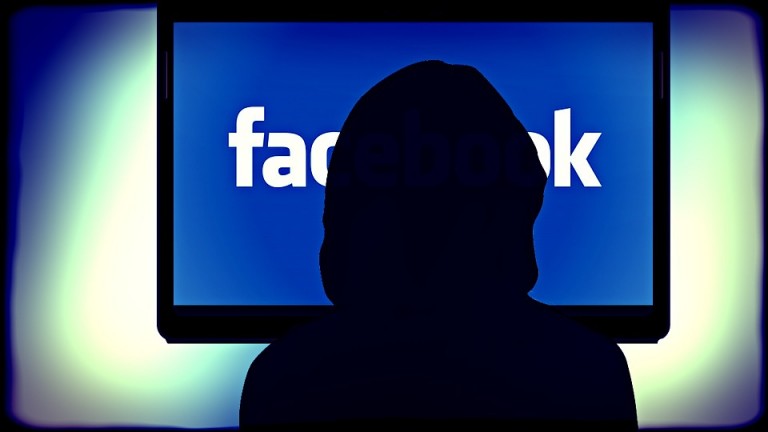 A figure wearing a black hoody in front of a screen with Facebook's logo on it