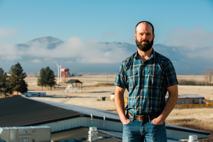 Photo of Jason standing on the roof of the Missoula Fire Lab with smoke jumper equipment behind him.