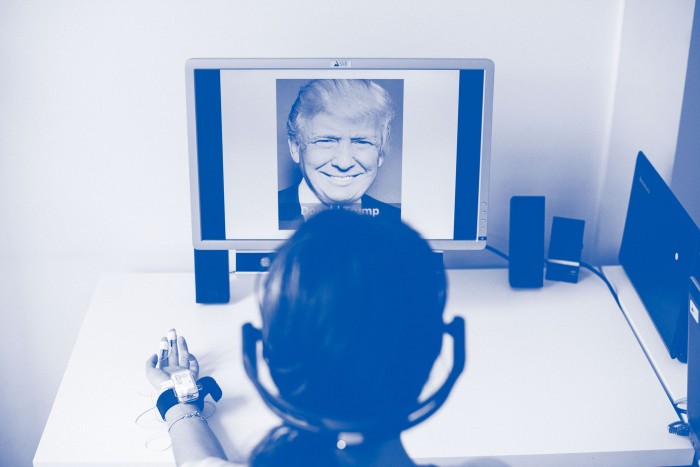 Photo of a woman wearing a monitor on her hand and headphones, looking at a computer screen displaying an image of Donald Trump.