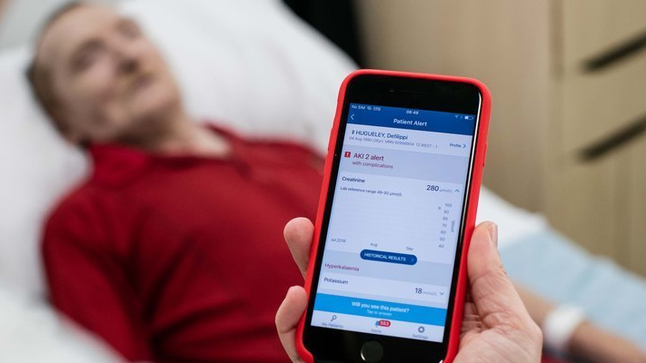 A man in a red polo top lying in a hospital bed behind a doctor holding up a smartphone